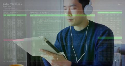 Asian man coding on tablet with headphones and stylus in home office, terminal data overlay