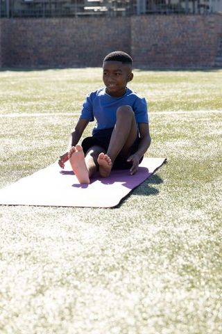 African american boy stretching on yoga mat outdoors on sunny school field