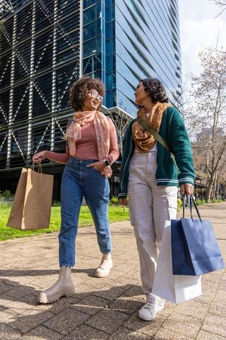 Two diverse women walking urban sidewalk carrying shopping bags laughing and chatting