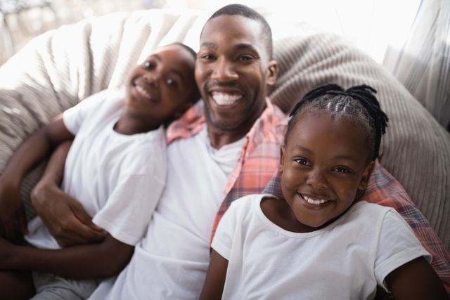 This image captures a joyful moment of a father with his daughter and son relaxing on a couch at home. The family is smiling and enjoying their time together, showcasing a strong bond and love. This image can be used for promoting family values, parenting, home life, and advertisements related to family-oriented products or services.