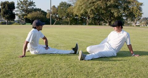 Cricket teammates relaxing on sunlit outfield during practice in white uniforms and black caps