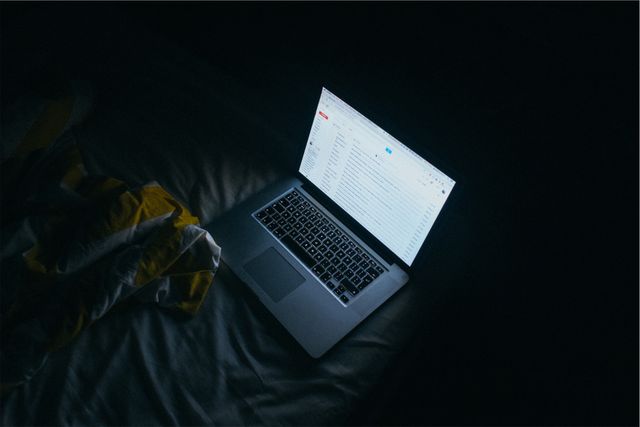 Laptop with illuminated screen showing an open email application placed on a bed in a dark room. Useful for illustrating concepts related to remote work, online communication, technology, working late, and nighttime productivity.