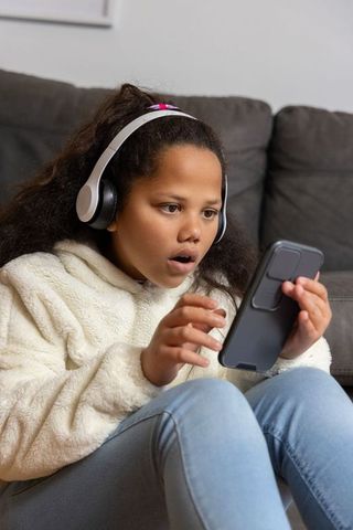 Surprised young girl wearing headphones holding smartphone on sofa reacting to video live