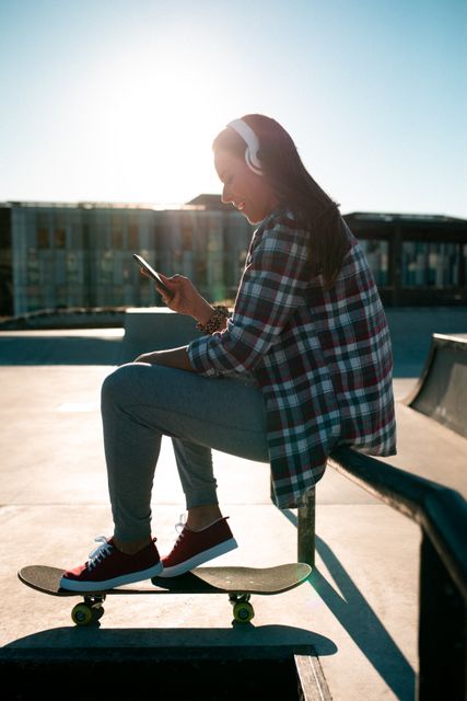 Woman enjoying a sunny day at an urban skatepark, wearing headphones and using her smartphone. Ideal for themes related to youth culture, technology, outdoor activities, and modern lifestyle. Perfect for promoting music streaming services, casual fashion brands, or urban lifestyle blogs.