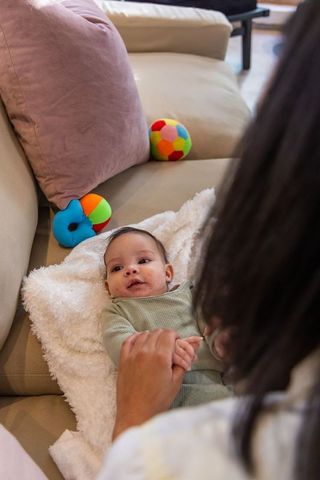 Caregiver holding infant hands while baby cuddling on beige sofa with plush blanket
