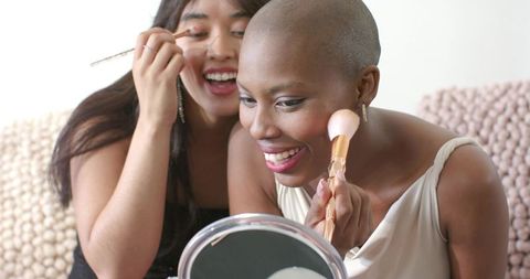 Smiling black and asian friends applying makeup together on cozy sofa using mirror and brushes