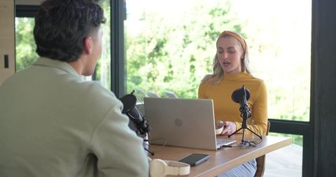 Podcast cohosts recording in bright home studio with laptop mics and greenery backdrop