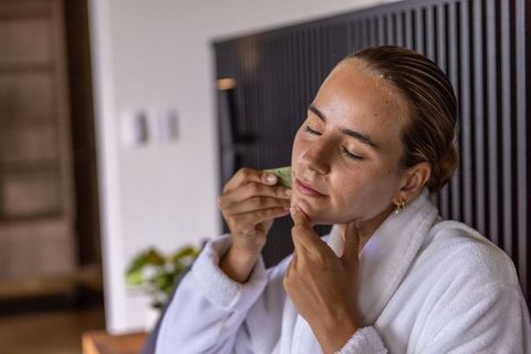 Woman in white robe applying green facial cloth for skincare and relaxation