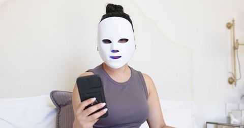 Woman wearing white led facial mask holding smartphone on bed for at-home skincare routine
