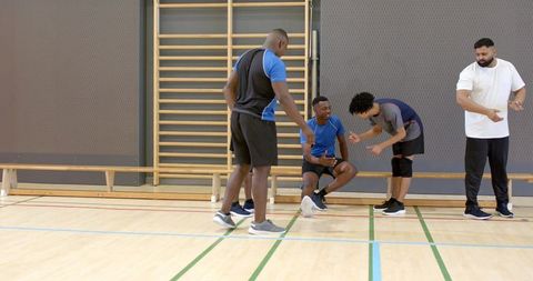 Diverse men supporting injured teammate during indoor basketball practice coach observing
