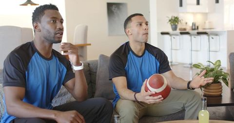 Friends watching football game on gray sofa holding ball reacting to big play at home