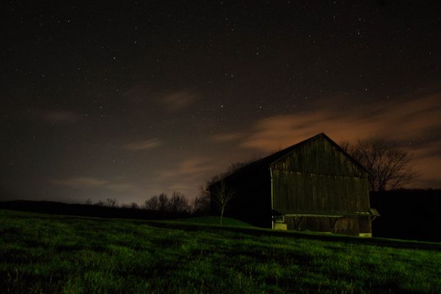 Beautiful rural scene with an old barn under a clear, starry night sky. The hint of a glow from the horizon suggests distant city lights. This image captures rural tranquility and the serene beauty of the countryside at night. Great for use in promotions related to agriculture, countryside living, calm and serene atmospheres.