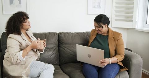 Women collaborating on laptop and drinking coffee on couch during casual home meeting