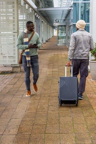 Two men walking with rolling suitcase and crossbody bag along modern glass corridor