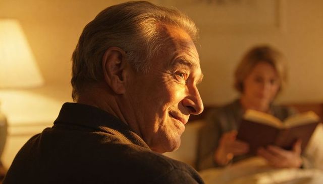 Senior man smiling in warm bedside light, partner reading hardcover book, cozy evening