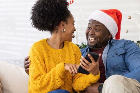 African american couple laughing and pointing at smartphone on couch wearing santa hat