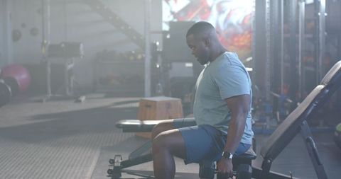 African american man sitting on adjustable bench holding dumbbells, backlit gym training