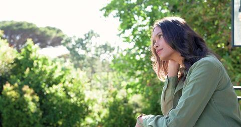 African woman gazing contemplatively from balcony into sunlit garden with olive jacket