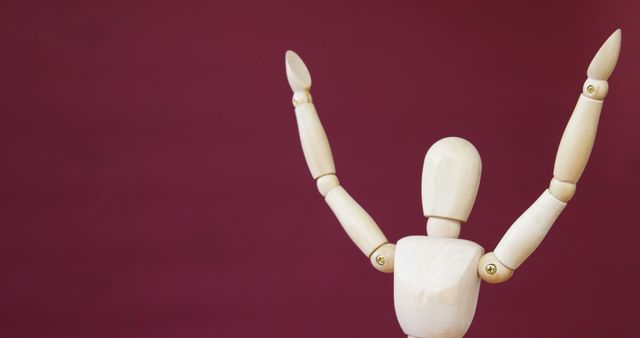 A wooden artist's mannequin is posed with arms raised against a maroon background, with copy space. Its neutral form and simple design make it a versatile tool for artists to study human movement and poses.