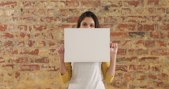 Portrait of happy biracial woman with long hair holding white board with copy space by brick wall. Lifestyle, mock up, message and information, unaltered.