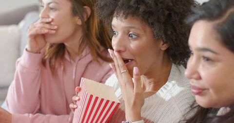 Diverse women enjoying movie night and reacting while sharing striped popcorn on couch