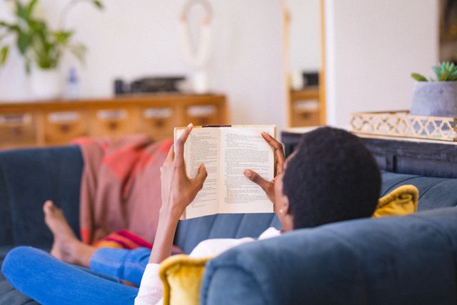 African american woman lying on sofa while reading novel and relaxing in living room at home. unaltered, people, lifestyle, home and hobbies concept.