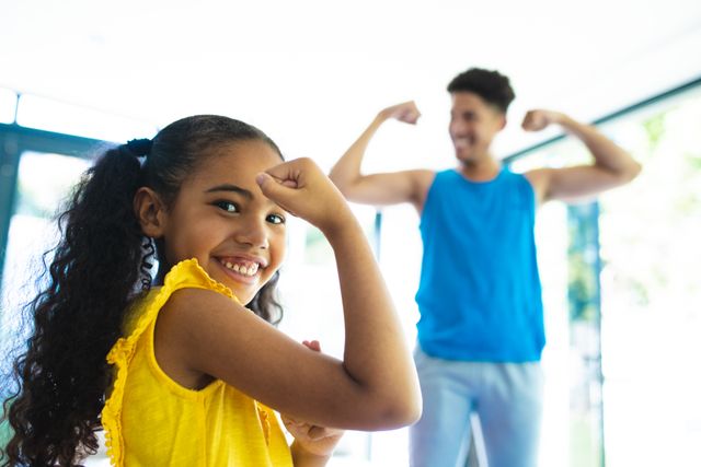 This image captures a joyful moment between a father and his daughter as they flex their muscles together at home. Perfect for use in family-oriented content, parenting blogs, fitness and health promotions, and lifestyle articles emphasizing family bonding and strength.