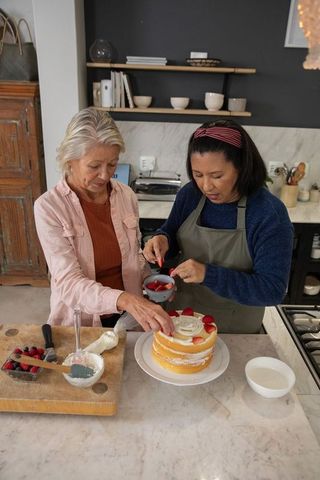 Two friends decorating layered strawberry cake on marble kitchen island, home baking