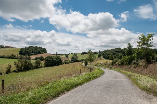 Rural country road winding through lush green landscape under a bright blue sky with fluffy clouds. Ideal for depicting peaceful rural life, traveling, journey, summertime outings and eco-tourism.
