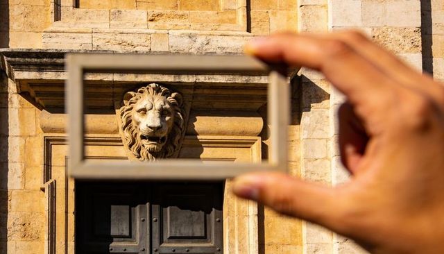 Hand framing carved lion head relief on sunlit historic limestone facade with doorway