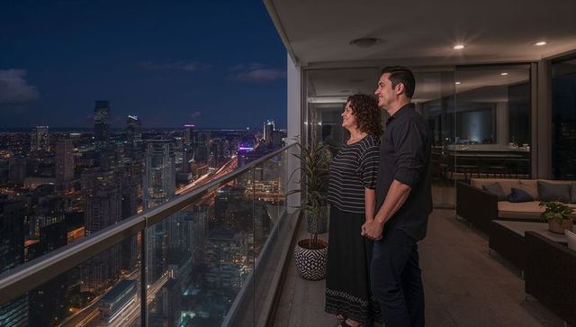 Holding hands couple gazing over night urban city skyline from luxury high-rise balcony