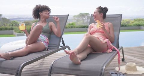 African american friends relaxing poolside on lounge chairs holding cocktails and laughing