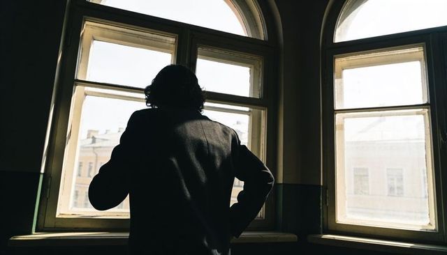 Silhouetted man standing by arched windows watching sunlit urban facades in dim interior