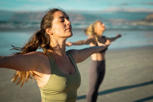 Two women practicing yoga on a beach during sunset. They are standing with arms outstretched, eyes closed, embracing the peaceful environment. Ideal for promoting outdoor fitness, healthy lifestyle, and mindfulness. Suitable for wellness blogs, fitness programs, and relaxation retreats.