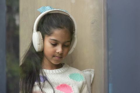 Young girl listening with white headphones in cozy window nook wearing knit sweater and blue bow