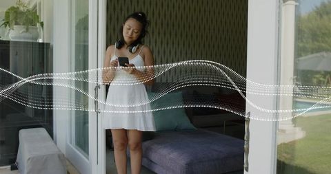 Woman standing in doorway wearing white dress and headphones checking phone with audio waveform