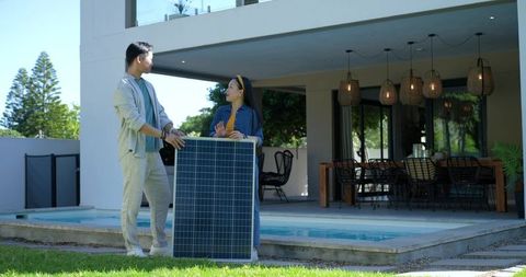 Asian couple installing large solar panel by poolside at modern home, discussing setup