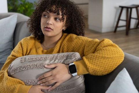 Contemplative african american woman on sofa holding pillow in mustard sweater with smartwatch