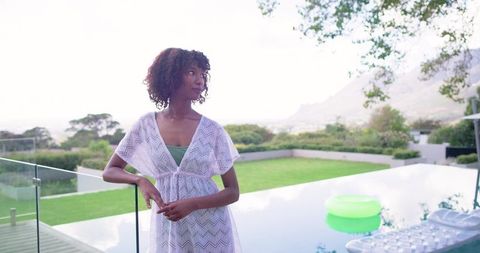 African woman leaning on glass balustrade by infinity pool wearing white cover-up gazing