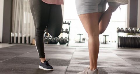 Women stretching single-leg balance in modern gym with dumbbell racks and backlit windows