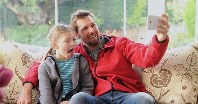 Father and daughter sit on the couch and take a playful selfie together. Warm and lighthearted moment showcases family bonding and fun at home. Perfect for themes related to family, parenting, smartphone usage, and leisure time. This could be used in advertisements, blogs, articles, and promotions focused on family life and technology.