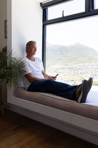 Mature man lounging on window bench with scenic mountain view holding smartphone