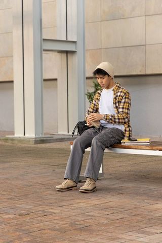 Asian teen sitting on bench in urban courtyard wearing beanie holding snack backpack