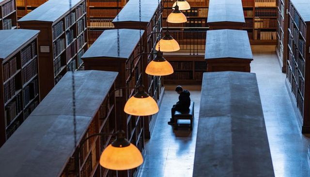 Student sitting under amber pendant lamps reading book in grand wooden library hall
