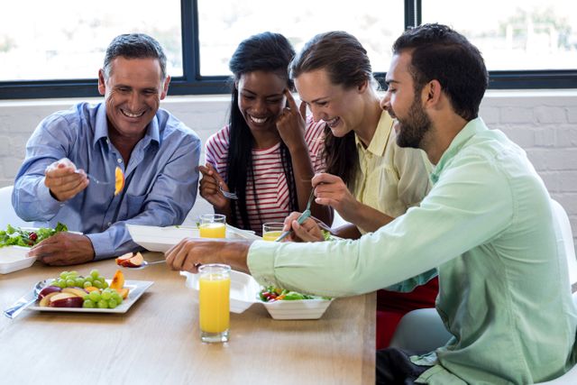Diverse group of colleagues enjoying breakfast together in an office setting. They are smiling and engaging in conversation while eating healthy meals and drinking orange juice. This image can be used to depict teamwork, office culture, professional bonding, and the importance of taking breaks together in a workplace environment.