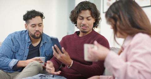 Sharing smartphone and coffee on cozy sofa, diverse young adults enjoying casual catch-up