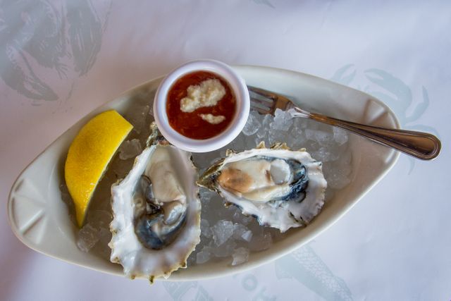 Image showing a gourmet dish with fresh oysters resting on ice, accompanied by a lemon wedge and a small dish of cocktail sauce, and a metal fork. This image is perfect for use in culinary blogs, seafood restaurant menus, food-related advertisements, and for highlighting gourmet dining experiences.