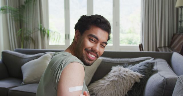 A young man smiling and showing a bandage on his arm while sitting in a living room with neutral-toned decor and large windows. This can be used to illustrate themes of health, vaccination, recovery, and home comfort.