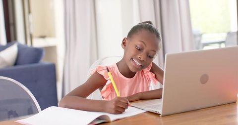 Smiling African American girl studying at laptop writing in notebook on home desk