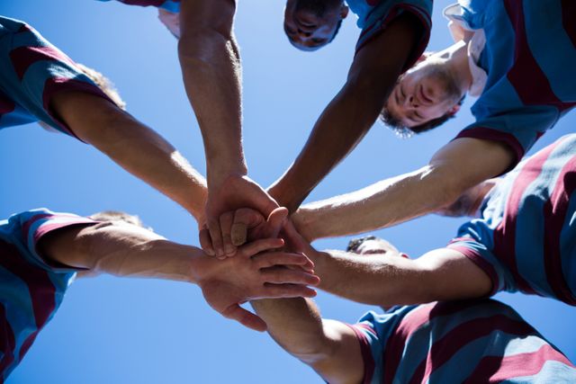 This image captures a rugby team showing unity and teamwork by holding hands in a huddle against a clear sky. Ideal for use in sports-related content, motivational materials, team-building promotions, and advertisements emphasizing cooperation and camaraderie.
